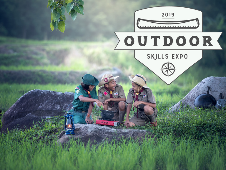 Children in outdoor uniforms cooking over a campstove in the middle of a meadow.